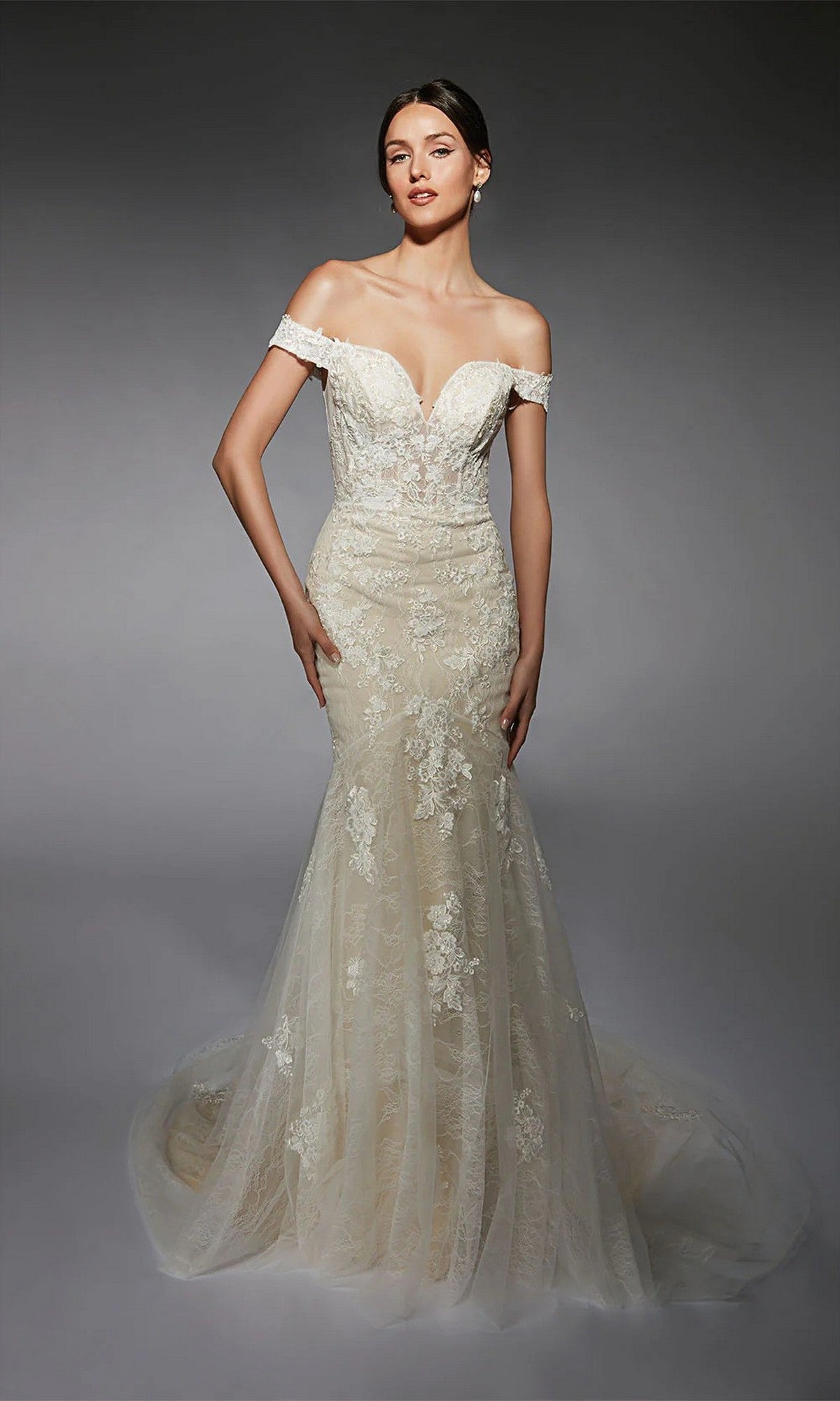 A woman wears the Alyce 7118 long white dress with an off-the-shoulder lace corset, sweetheart neckline, and long train, standing against a dark background.